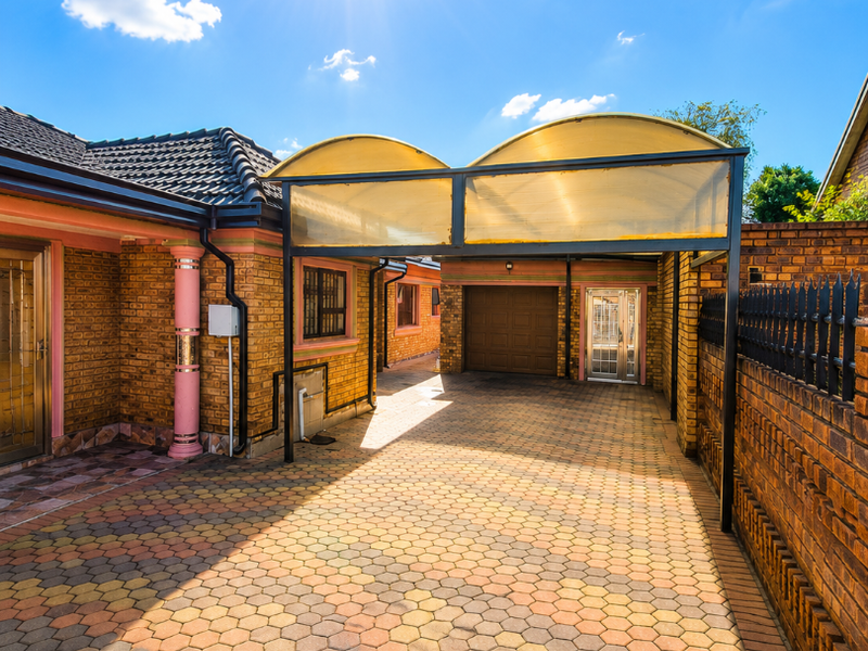 A well-maintained front exterior featuring brick paving, a covered carport, and a secure brick boundary wall with a garage entrance.