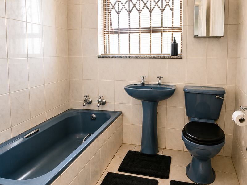 A functional bathroom featuring a blue bathtub, pedestal basin, and toilet, finished with neutral wall and floor tiles and a security-fitted window.