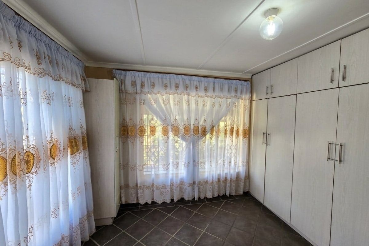 A bedroom featuring built-in cupboards, tiled flooring, and large windows with curtains, offering a clean and functional space.