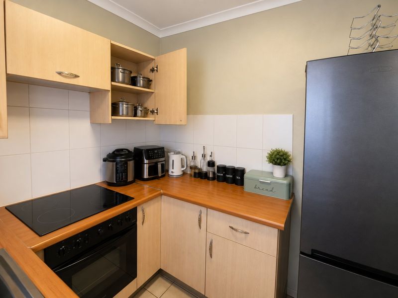 A functional kitchen featuring light wood-finish cabinetry, a laminate countertop, a built-in electric hob and oven, and white tiled splashbacks.