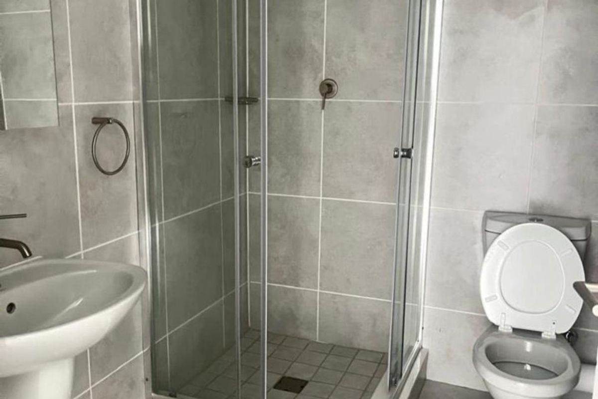 A clean, modern bathroom featuring a glass shower enclosure, pedestal basin, and toilet with neutral grey wall and floor tiling.