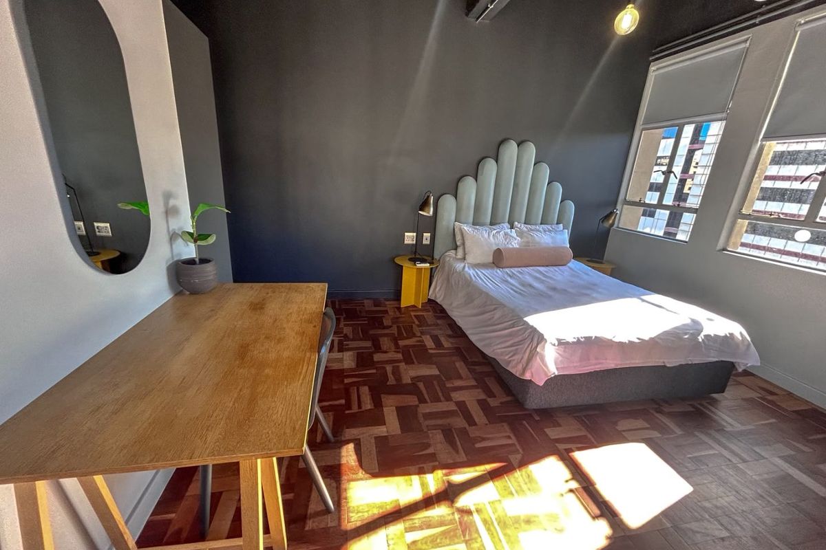 A well-lit bedroom featuring classic parquet flooring, dark accent walls, and large windows with roller blinds.