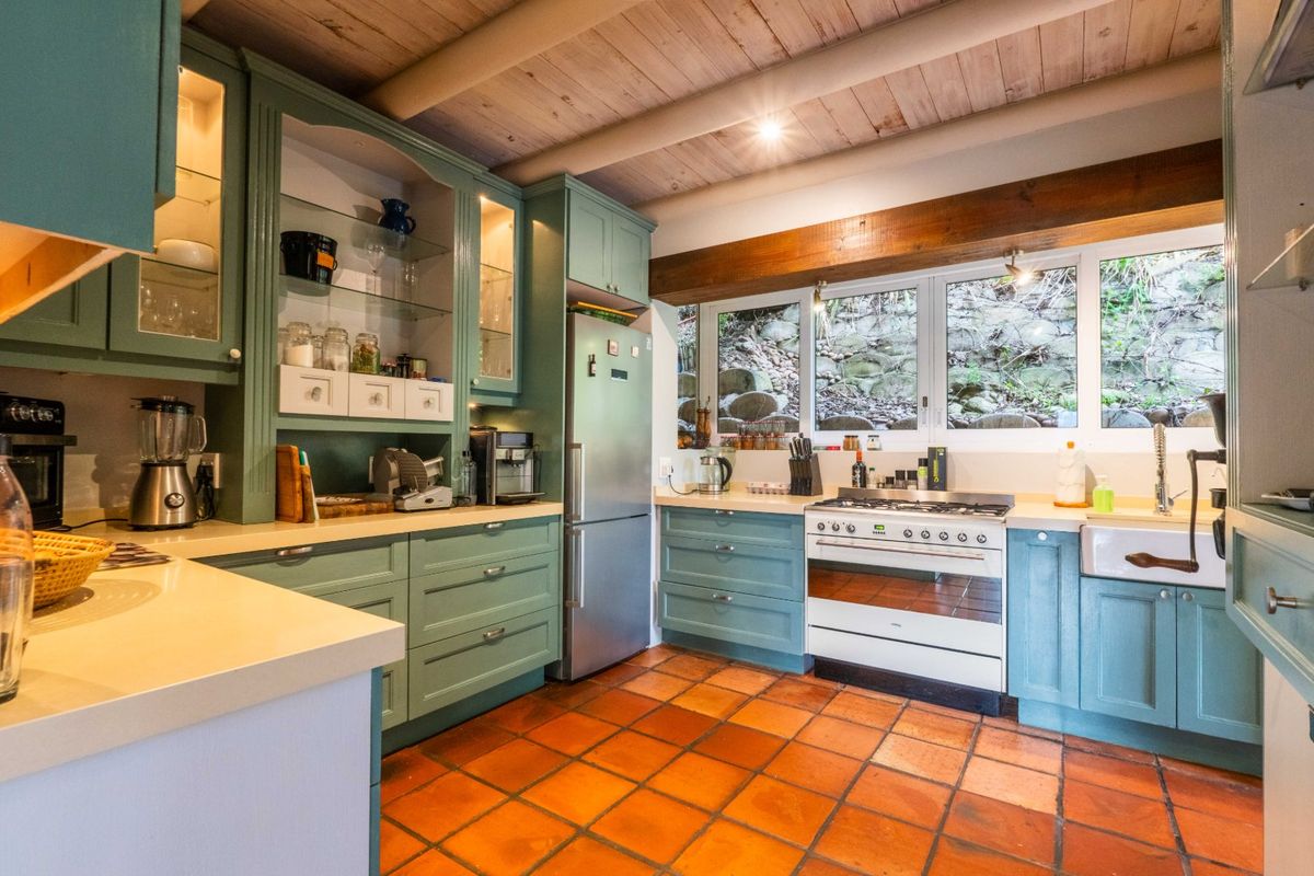 A charming kitchen featuring sage green cabinetry, terracotta floor tiles, exposed timber ceiling beams, and a classic apron sink with views of a stone wall.