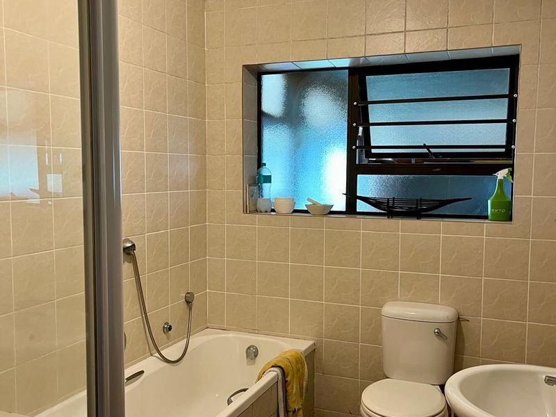 A functional bathroom featuring a bathtub with shower screen, pedestal basin, toilet, and neutral wall tiling with a louver window.