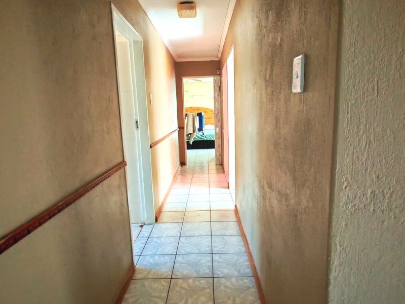 A narrow residential hallway featuring tiled flooring and a chair rail, leading towards a bedroom entrance.