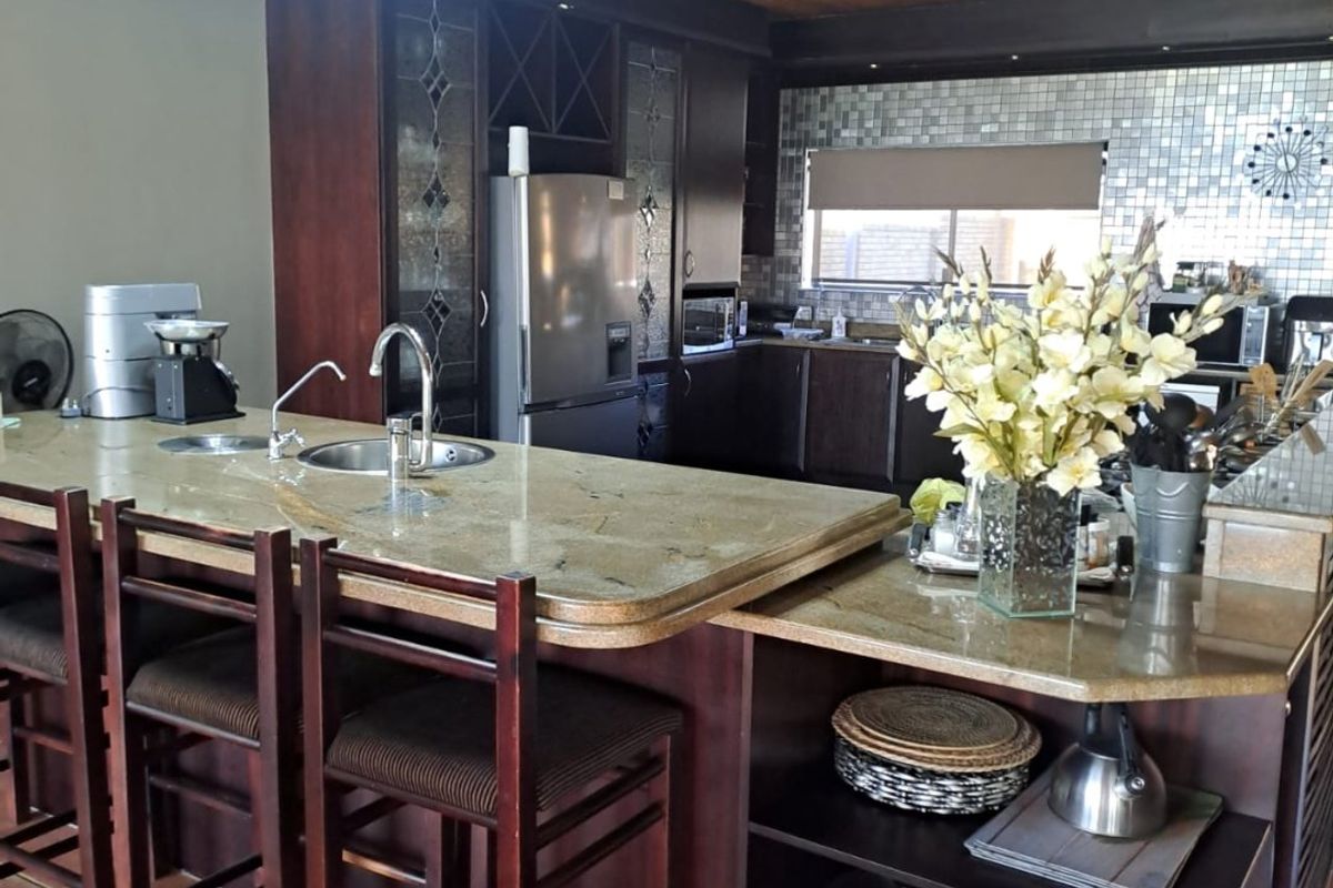 A spacious kitchen featuring a large granite-topped island, dark wood cabinetry, and a distinctive timber ceiling with mosaic tile backsplash detailing.