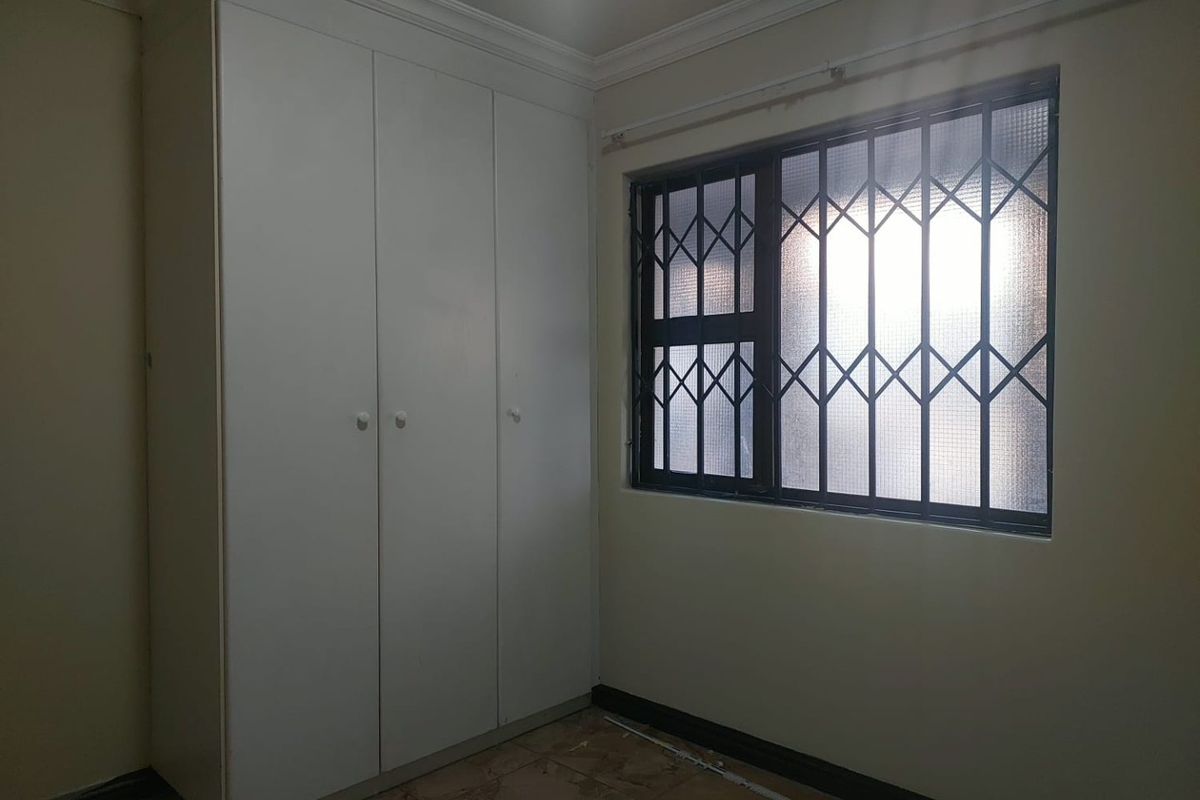 A vacant bedroom featuring built-in white wardrobes, tiled flooring, and a window fitted with a security gate.