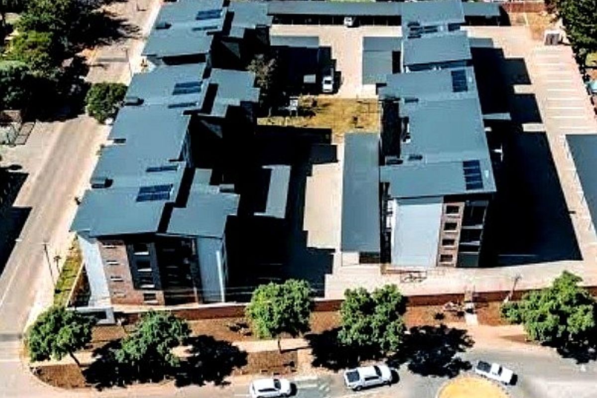 Aerial view of a multi-story apartment complex featuring pitched roofs with solar panels, paved parking, and surrounding landscaped grounds.