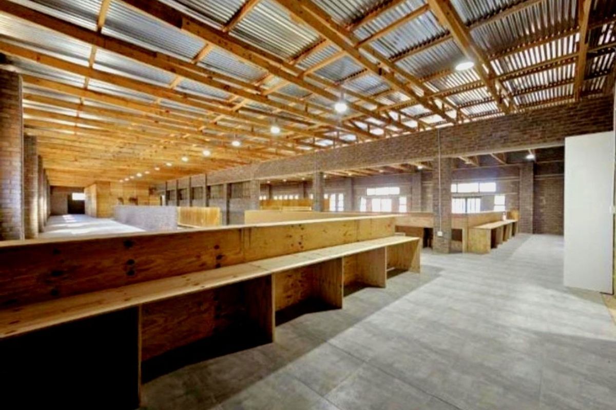 Spacious open-plan commercial interior featuring exposed timber roof trusses, corrugated metal ceiling, exposed brickwork, and built-in plywood counters on tiled flooring.
