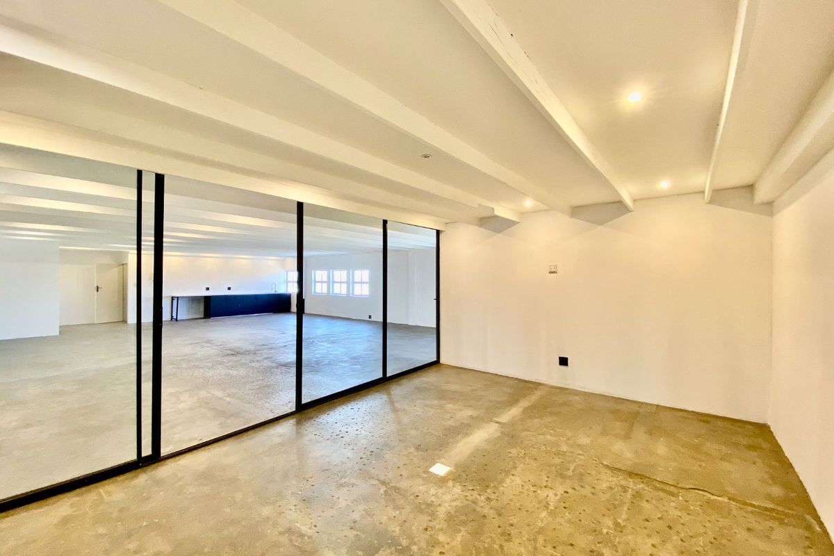 Spacious open-plan commercial area featuring polished concrete floors, exposed ceiling beams, and a modern black-framed glass partition separating the main space from the kitchen.