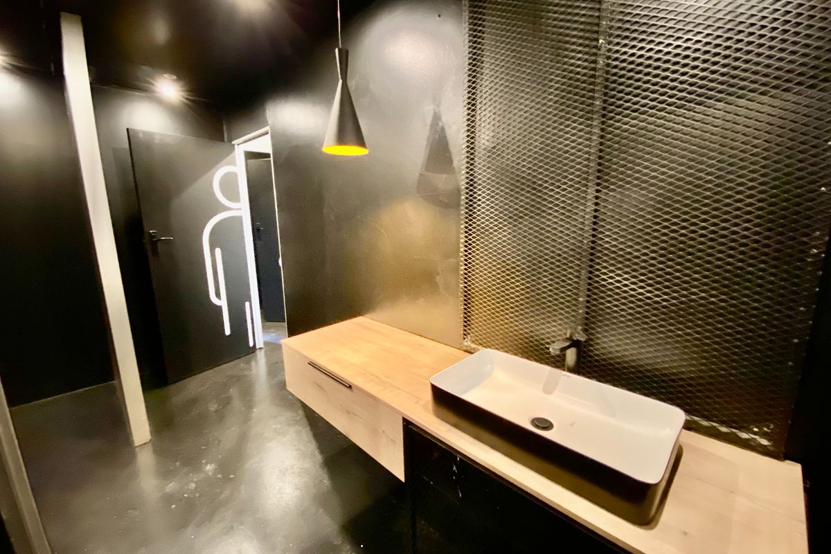 Modern commercial bathroom featuring a floating wooden vanity, vessel sink, and industrial-style metal mesh wall accents with dark contemporary finishes.
