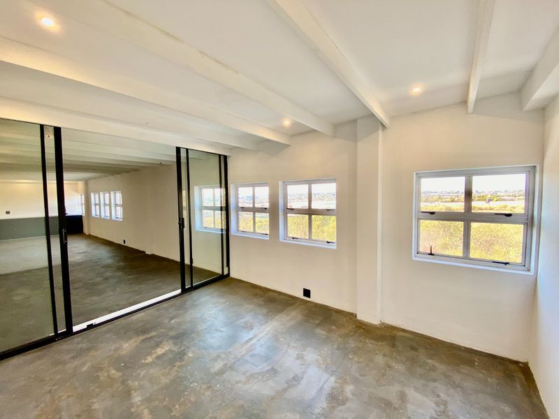 A bright, open-plan office space featuring exposed beam ceilings, recessed lighting, concrete flooring, and a modern sliding glass partition with scenic exterior views.