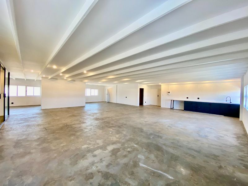 Spacious open-plan commercial area featuring exposed beam ceilings, recessed lighting, and a functional kitchenette corner with a sink and built-in cabinetry.