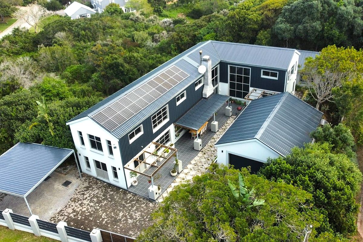Aerial view of a modern multi-level home featuring a metal roof, integrated solar panels, a paved courtyard with a pergola, and a detached garage.