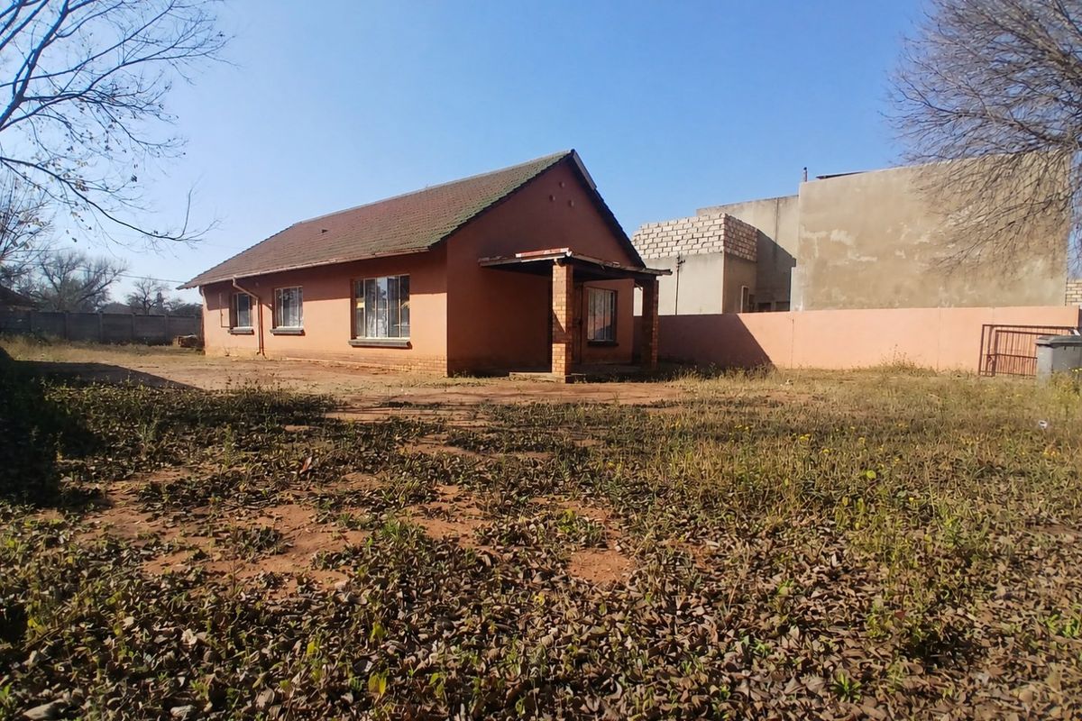 Rear view of a detached house featuring a tiled roof, brick facade, and a covered porch, set on a large plot with unmaintained grounds.