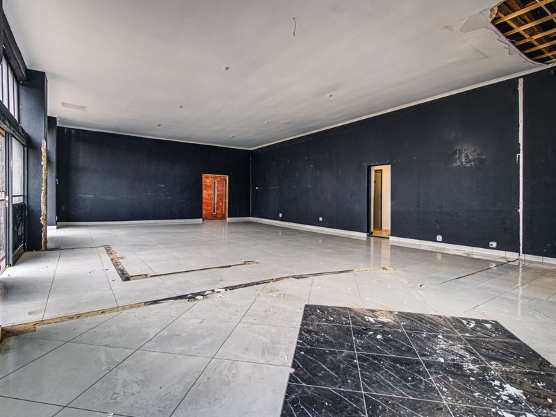 A large, vacant commercial open-plan space featuring tiled flooring, large storefront windows, and significant ceiling and flooring repairs required.