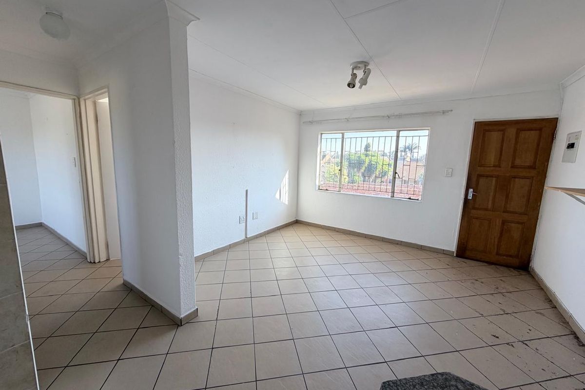 An empty, open-plan living area featuring neutral tiled flooring, security bars on the window, and a classic timber door.