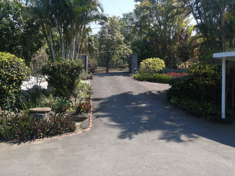 A private driveway leading to a metal security gate, surrounded by established trees and landscaped garden beds with brick edging.