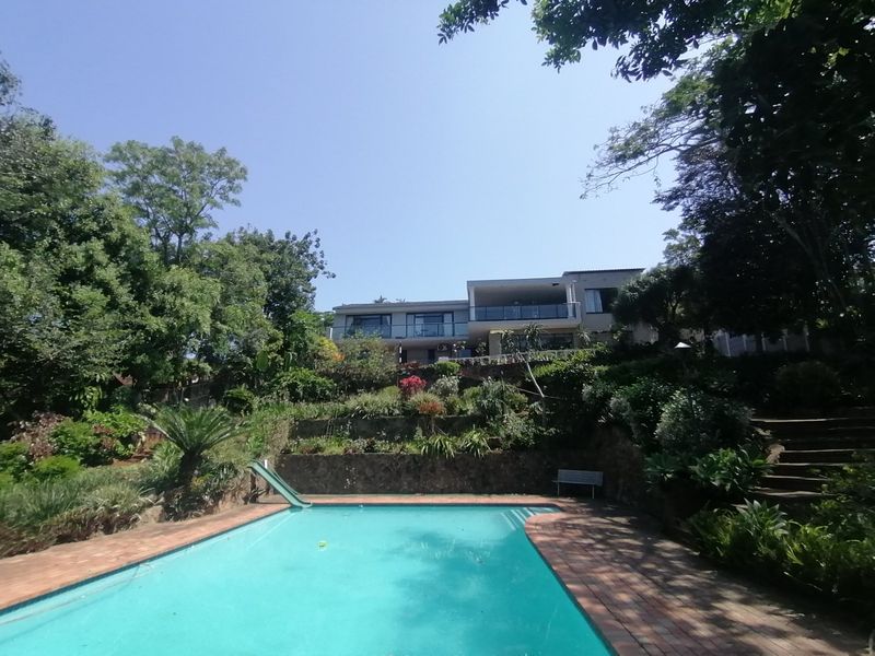 A spacious outdoor pool area featuring a swimming pool with a slide, surrounded by tiered landscaped gardens and a stone retaining wall.