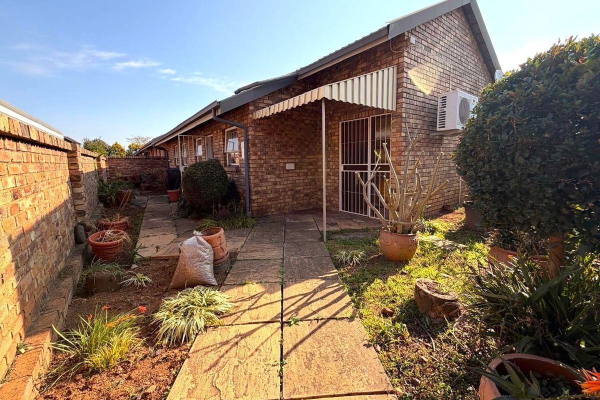 Rear exterior view of a facebrick townhouse featuring a paved walkway, security gate, and private garden area with an installed air conditioning unit.