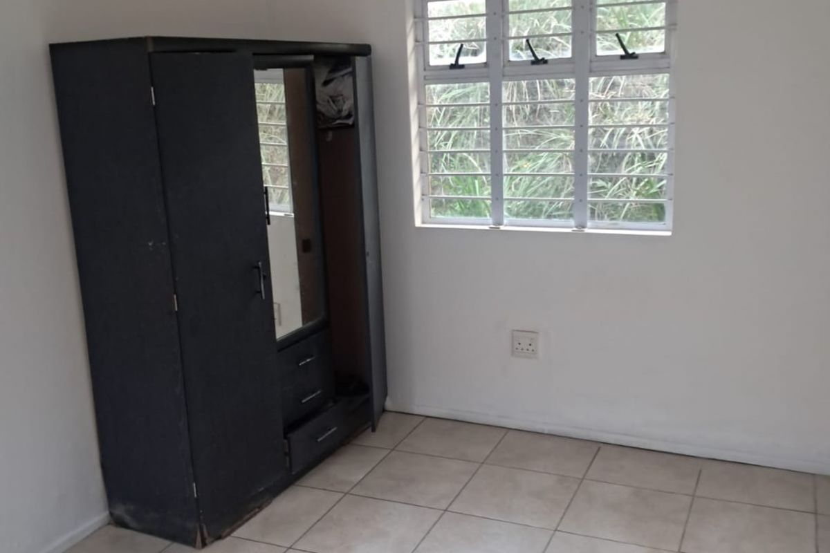 A simple bedroom featuring tiled flooring, a window with security bars, and neutral wall finishes, offering a functional space ready for personalization.