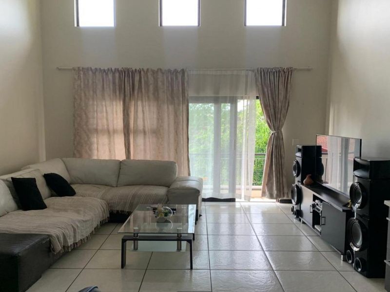 Spacious open-plan living area featuring double-volume ceilings, large windows for natural light, tiled flooring, and direct access to a balcony.