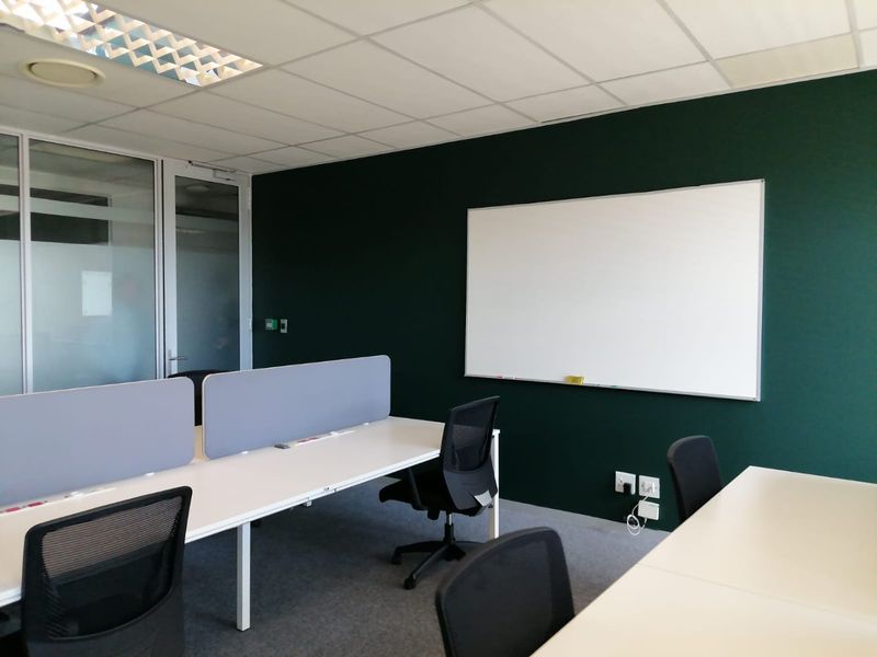A modern office space featuring a dark green accent wall, a mounted whiteboard, glass partitioning, and a suspended ceiling with recessed lighting.