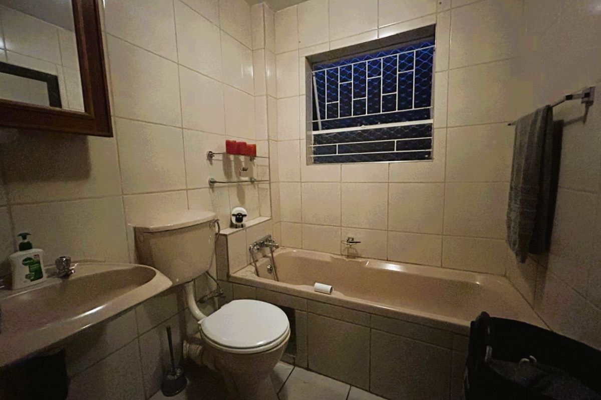 A dated bathroom featuring a bathtub, toilet, and pedestal basin with full-height wall tiling and a window with security bars.