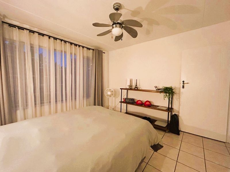 A bedroom featuring tiled flooring, a ceiling fan, and large windows with curtain tracks, providing a functional and well-lit living space.