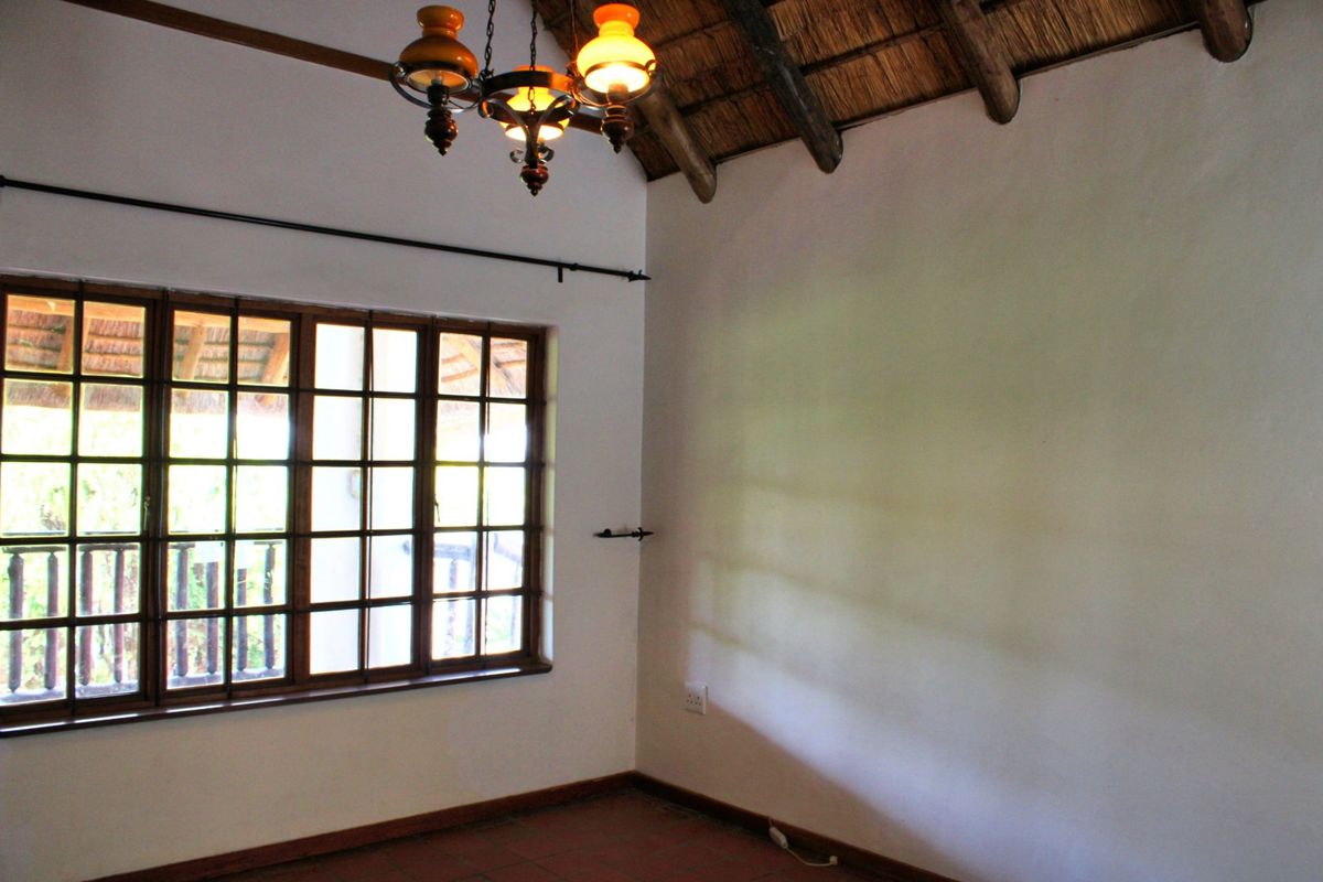 A bright, empty bedroom featuring a traditional thatched roof with exposed timber rafters, tiled flooring, and large timber-framed windows.