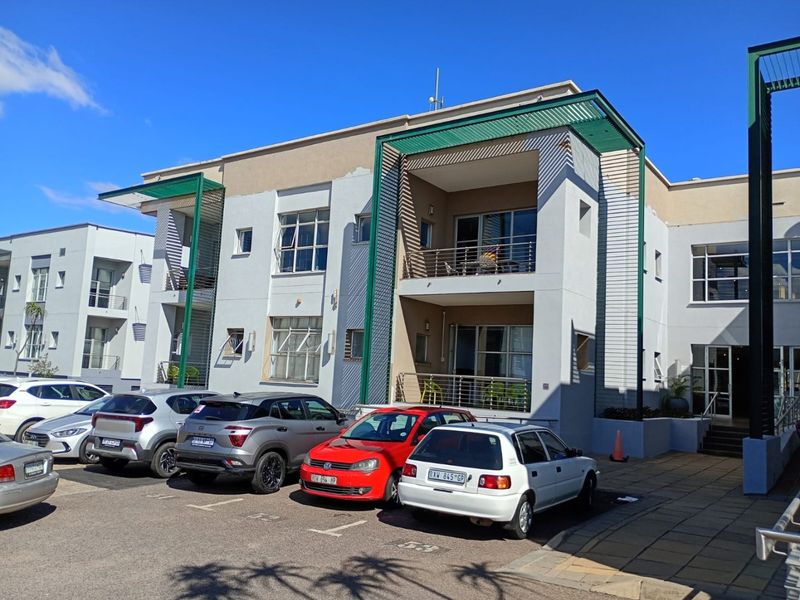 Front elevation of a commercial building featuring multi-level balconies with metal railings, green architectural pergolas, and an adjacent paved parking area.