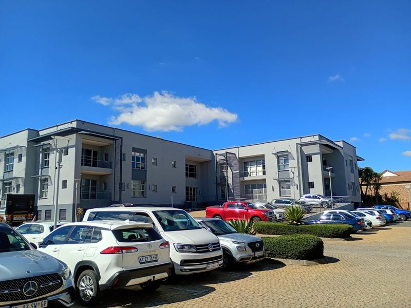 A modern multi-story commercial office building featuring a paved parking area, landscaped hedges, and private balconies for occupants.