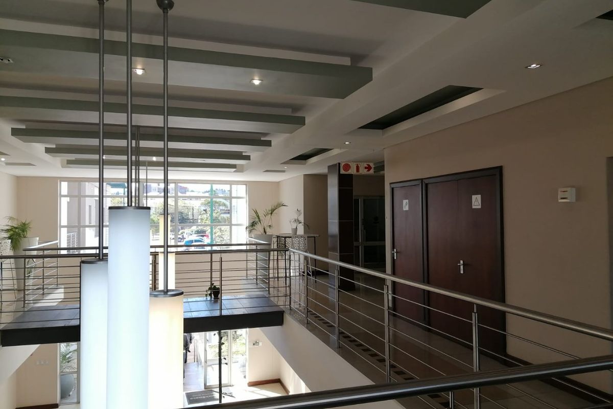 A spacious commercial hallway featuring modern stainless steel balustrades, recessed ceiling lighting, and clean tiled flooring with access to office facilities.