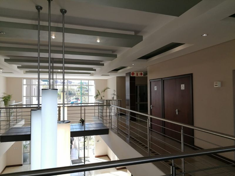 A spacious commercial hallway featuring modern stainless steel balustrades, recessed ceiling lighting, and clean tiled flooring with access to office facilities.