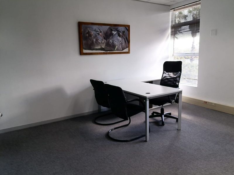 A functional office space featuring carpeted flooring, a suspended ceiling, and natural light provided by a window with venetian blinds.