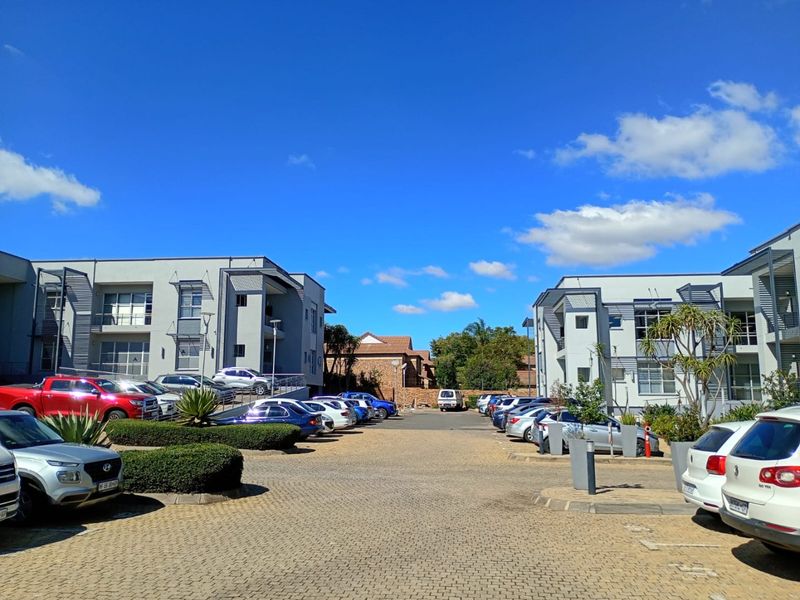 Exterior view of a modern commercial office complex featuring paved parking areas, landscaped garden beds, and multi-story building facades under a clear sky.