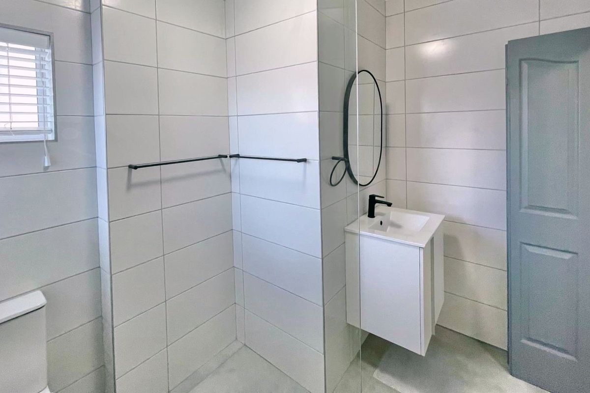 A modern, clean bathroom featuring white wall tiles, a wall-mounted vanity with a black mixer tap, and a glass shower enclosure.