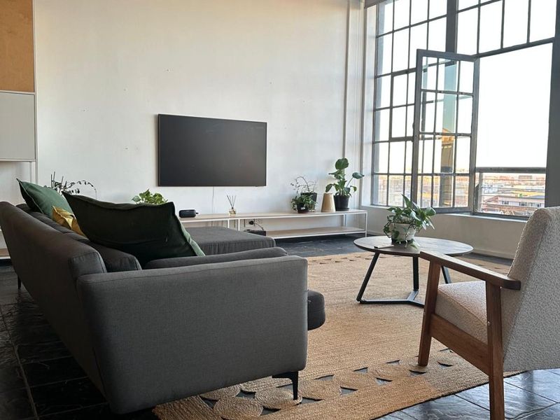 Spacious industrial-style living room featuring high ceilings with exposed ducting, large steel-framed windows, and dark stone tile flooring.