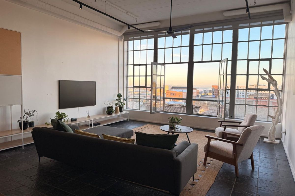 Spacious industrial-style living room featuring large steel-framed windows, exposed ceiling ducting, track lighting, and dark stone tile flooring with an urban view.