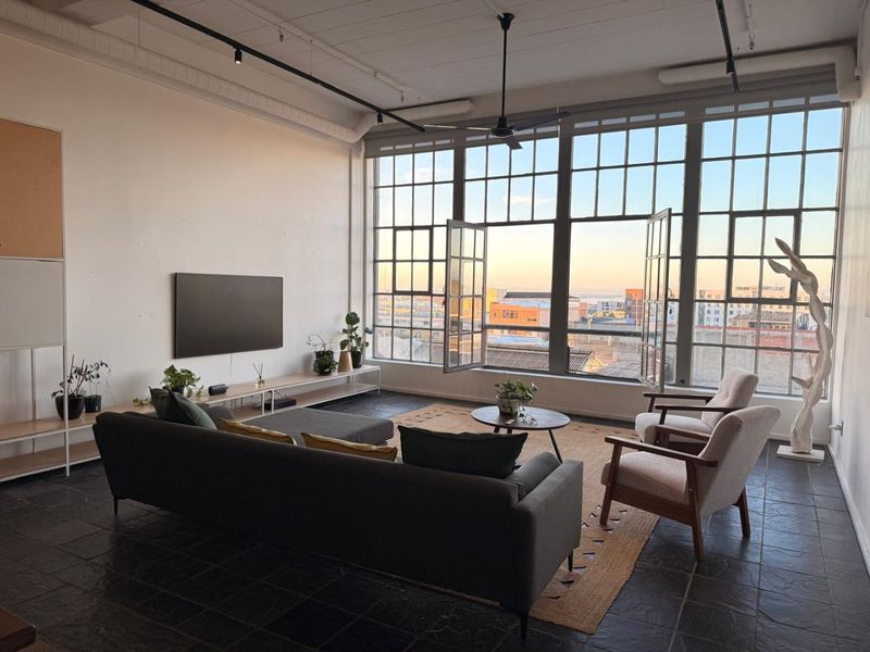 Spacious industrial-style living room featuring large steel-framed windows, exposed ceiling ducting, track lighting, and dark stone tile flooring with an urban view.