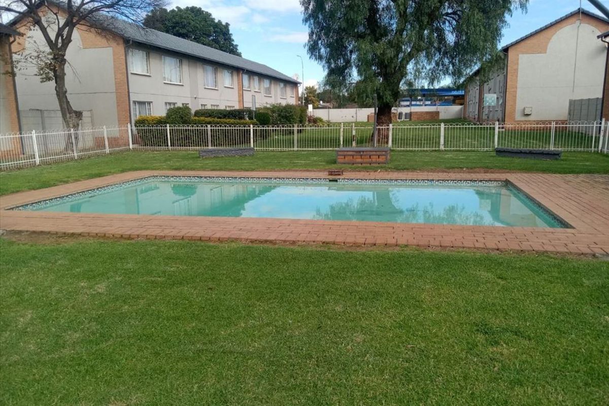 A communal swimming pool area featuring a brick-edged pool, surrounding lawn, and white metal fencing within an apartment complex setting.
