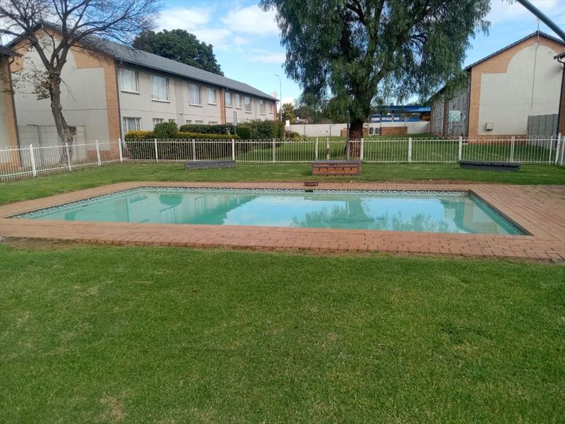 A communal swimming pool area featuring a brick-edged pool, surrounding lawn, and white metal fencing within an apartment complex setting.