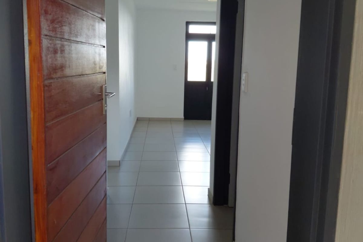 A bright, tiled hallway featuring a wooden door and clean white walls, leading towards an exterior doorway.