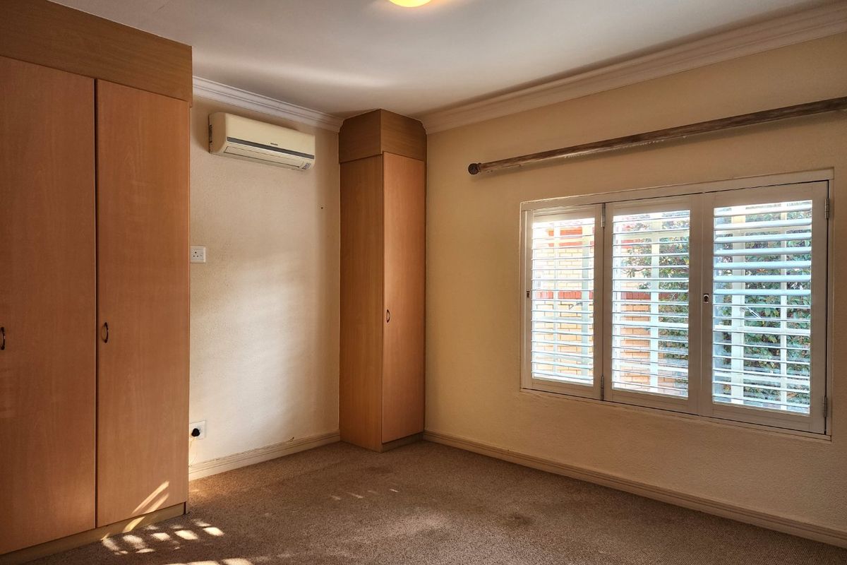 A vacant bedroom featuring built-in wooden cupboards, a wall-mounted air conditioning unit, and window shutters, providing a functional and neutral living space.