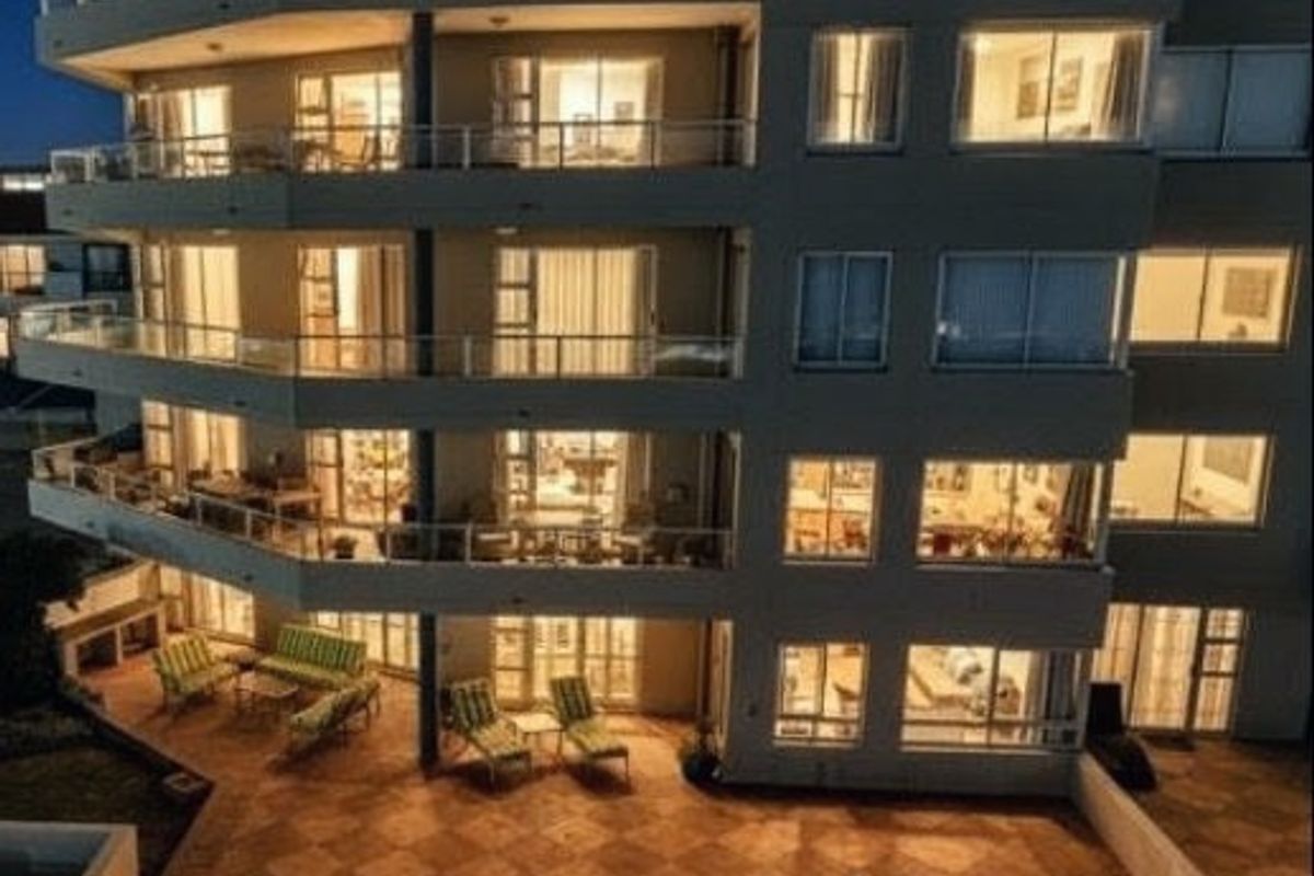 Evening view of an apartment building facade featuring multiple levels of balconies with glass balustrades and a ground-level tiled patio area.