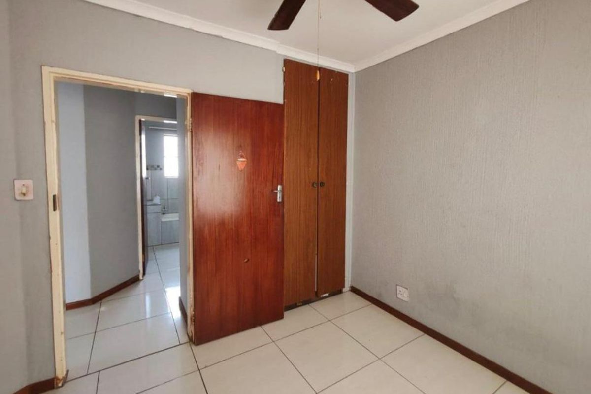 A vacant bedroom featuring tiled flooring, built-in wooden cupboards, and a ceiling fan, with an adjacent view into a bathroom area.
