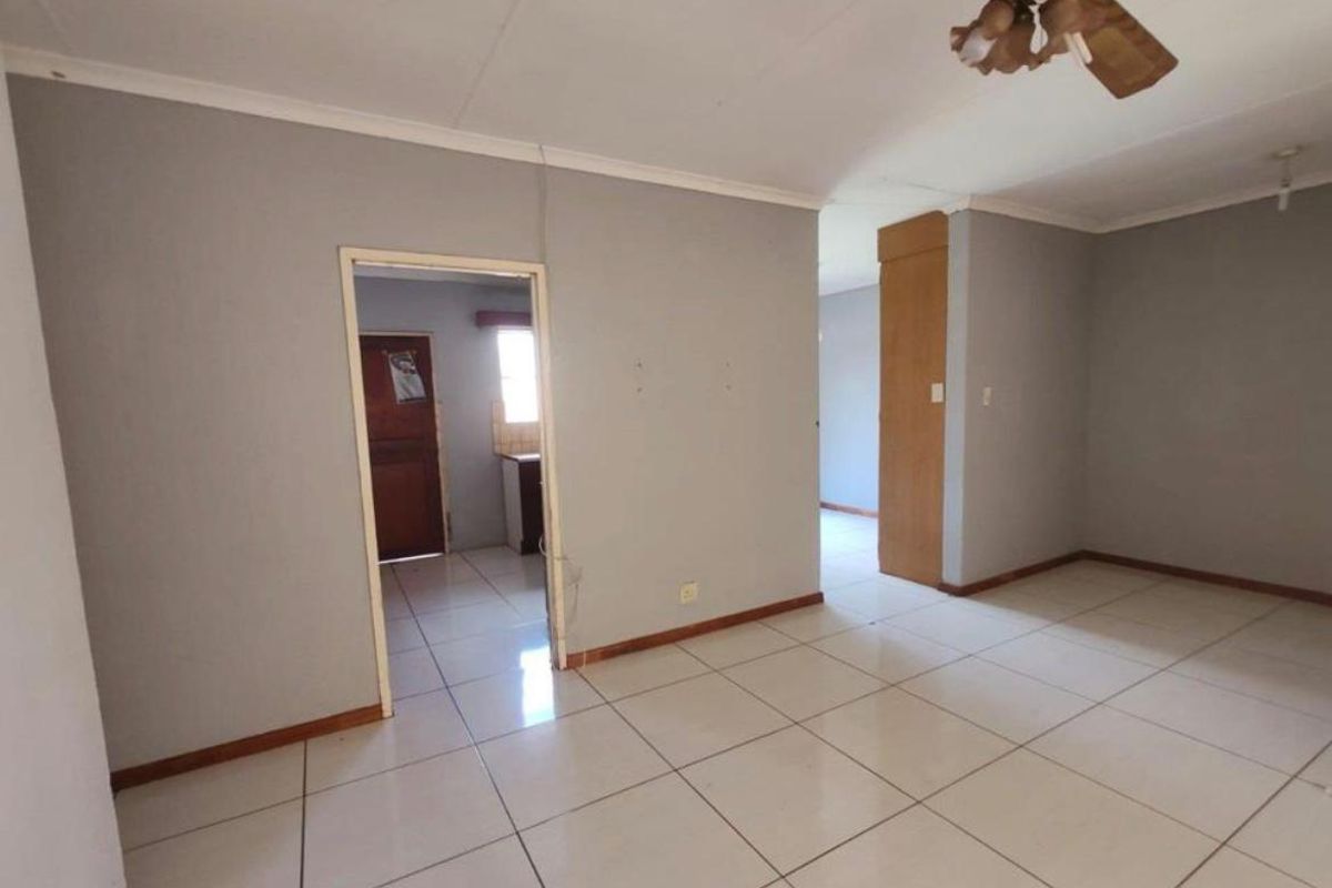 An empty living room featuring tiled flooring, coving, and a ceiling fan, with access to an adjacent kitchen area.