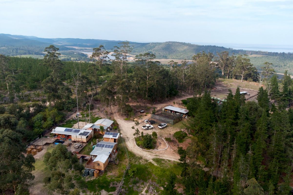 Aerial view of a rural farm property featuring multiple structures with solar installations, water tanks, and surrounding forest landscape.