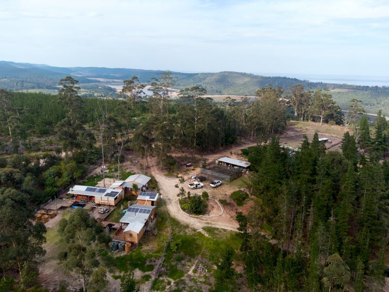 Aerial view of a rural farm property featuring multiple structures with solar installations, water tanks, and surrounding forest landscape.