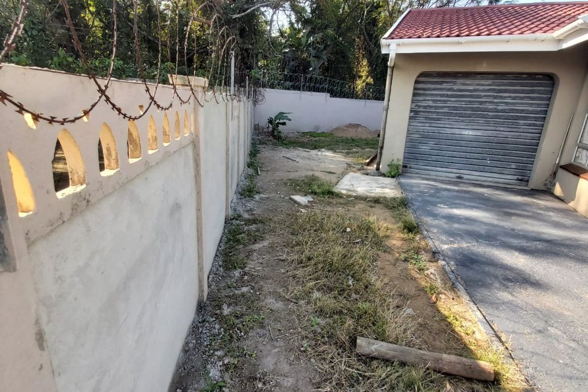 Rear exterior view showing a garage, paved driveway, and perimeter wall with security razor wire, featuring an unmaintained garden area.